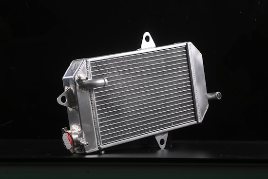 A metallic car radiator is placed against a dark background, highlighting its shiny silver surface and intricate grid of cooling fins. The radiator has several metal connectors protruding from its sides, and a cap with a caution label is visible in the lower area.