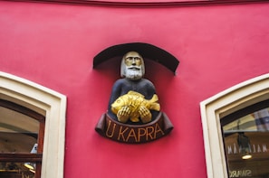 A decorative sign featuring a bearded figure holding a gold fish, mounted on a vividly painted pink wall. The sign has an arch above it and the words 'U Kapra' written below in golden letters.