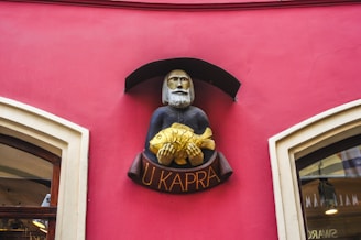 A decorative sign featuring a bearded figure holding a gold fish, mounted on a vividly painted pink wall. The sign has an arch above it and the words 'U Kapra' written below in golden letters.