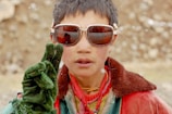 A young person wearing large sunglasses and holding up two gloved fingers in a peace sign. The individual is dressed in a mix of colorful clothing, including a red and green jacket and a yellow scarf. They have short, dark hair and appear to be outdoors, possibly in a snowy environment.
