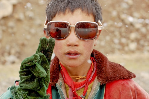 A young person wearing large sunglasses and holding up two gloved fingers in a peace sign. The individual is dressed in a mix of colorful clothing, including a red and green jacket and a yellow scarf. They have short, dark hair and appear to be outdoors, possibly in a snowy environment.
