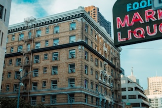 A multi-story brick building with numerous windows and fire escapes is situated in an urban environment. Nearby, a large neon sign displaying the words 'MARKET LIQUOR' is partially visible, with other buildings and skyscrapers in the background.