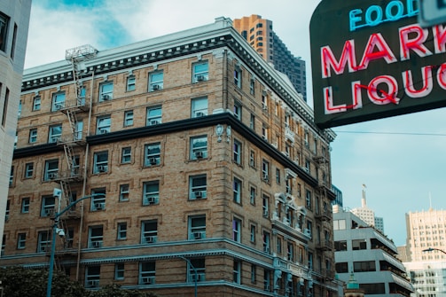 A multi-story brick building with numerous windows and fire escapes is situated in an urban environment. Nearby, a large neon sign displaying the words 'MARKET LIQUOR' is partially visible, with other buildings and skyscrapers in the background.