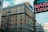 A multi-story brick building with numerous windows and fire escapes is situated in an urban environment. Nearby, a large neon sign displaying the words 'MARKET LIQUOR' is partially visible, with other buildings and skyscrapers in the background.