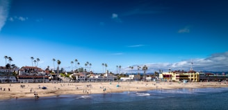 A bustling beach scene features numerous people enjoying various activities along the shoreline. The sandy beach is dotted with colorful umbrellas and beachgoers, with a backdrop of palm trees and charming coastal buildings. The vibrant blue sky is mostly clear, adding to the lively atmosphere.