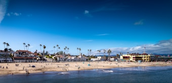 A bustling beach scene features numerous people enjoying various activities along the shoreline. The sandy beach is dotted with colorful umbrellas and beachgoers, with a backdrop of palm trees and charming coastal buildings. The vibrant blue sky is mostly clear, adding to the lively atmosphere.