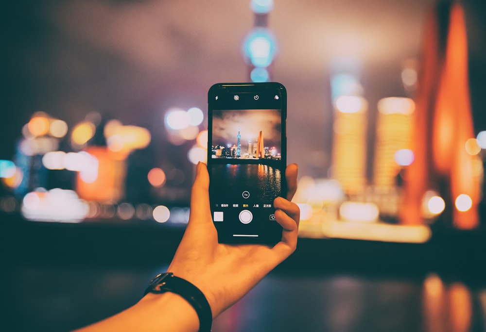 person holding phone taking photo of buildings at night