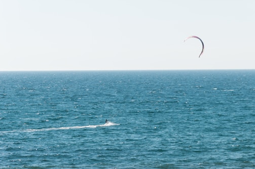 A lone kitesurfer navigates the expansive ocean waters with a visibly arched kite soaring in the sky. The horizon separates the deep blue sea from the pale blue sky, creating a serene and open atmosphere.