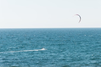 A lone kitesurfer navigates the expansive ocean waters with a visibly arched kite soaring in the sky. The horizon separates the deep blue sea from the pale blue sky, creating a serene and open atmosphere.