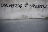 Graffiti on a concrete wall with the words 'LIBERATION OF ENSLAVED' incorporated into the text. The text is spray-painted in black with some letters using an anarchy symbol.