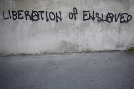 Graffiti on a concrete wall with the words 'LIBERATION OF ENSLAVED' incorporated into the text. The text is spray-painted in black with some letters using an anarchy symbol.