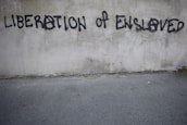 Graffiti on a concrete wall with the words 'LIBERATION OF ENSLAVED' incorporated into the text. The text is spray-painted in black with some letters using an anarchy symbol.