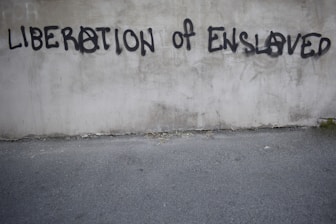 Graffiti on a concrete wall with the words 'LIBERATION OF ENSLAVED' incorporated into the text. The text is spray-painted in black with some letters using an anarchy symbol.