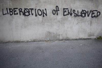 Graffiti on a concrete wall with the words 'LIBERATION OF ENSLAVED' incorporated into the text. The text is spray-painted in black with some letters using an anarchy symbol.