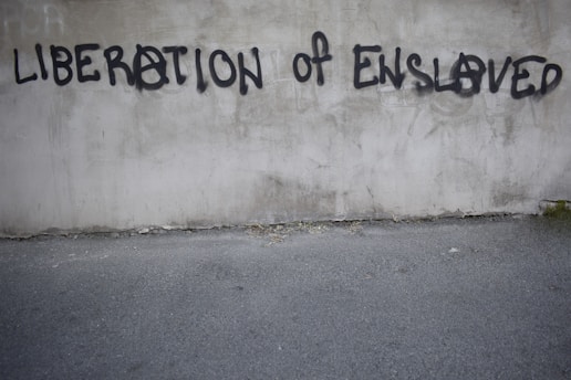 Graffiti on a concrete wall with the words 'LIBERATION OF ENSLAVED' incorporated into the text. The text is spray-painted in black with some letters using an anarchy symbol.