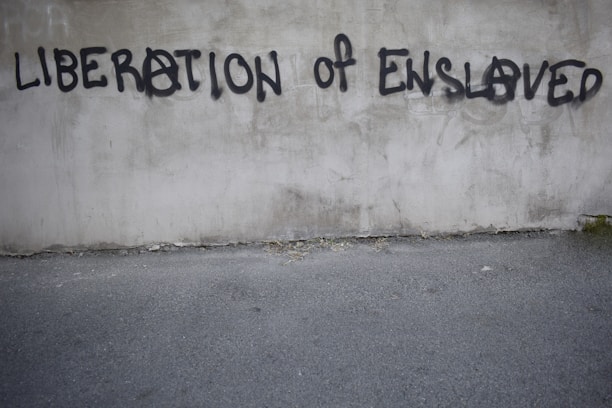 Graffiti on a concrete wall with the words 'LIBERATION OF ENSLAVED' incorporated into the text. The text is spray-painted in black with some letters using an anarchy symbol.
