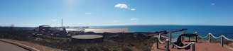 A coastal industrial site with various factory buildings and chimneys along the shore. The foreground features a fenced viewpoint with nautical decor, including ropes and an anchor. The ocean stretches out to the horizon under a clear blue sky.