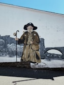 A mural painted on a wall features an elderly man in traditional, medieval attire, complete with a wide-brimmed hat and a staff. The background includes artistic renditions of stone architecture, trees, and a bridge, all in a grayscale color palette.