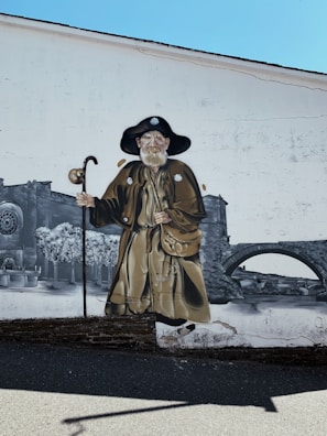 A mural painted on a wall features an elderly man in traditional, medieval attire, complete with a wide-brimmed hat and a staff. The background includes artistic renditions of stone architecture, trees, and a bridge, all in a grayscale color palette.