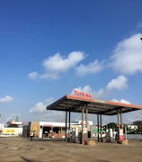 A gas station with the Total brand under a clear blue sky with scattered clouds. The station has multiple fuel pumps and a convenience store with signage saying 'bonjour'. There are paved paths and parking lanes in front of the station.
