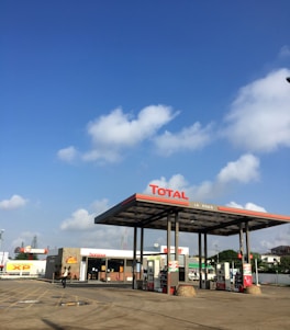 A gas station with the Total brand under a clear blue sky with scattered clouds. The station has multiple fuel pumps and a convenience store with signage saying 'bonjour'. There are paved paths and parking lanes in front of the station.
