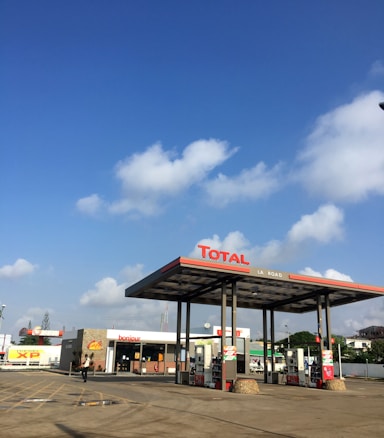 A gas station with the Total brand under a clear blue sky with scattered clouds. The station has multiple fuel pumps and a convenience store with signage saying 'bonjour'. There are paved paths and parking lanes in front of the station.