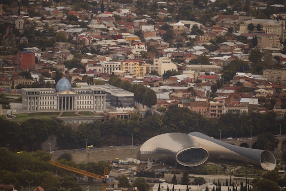 An aerial view of a cityscape featuring a large, domed government building surrounded by multiple smaller residential structures. In the foreground, there is a unique, modern architectural structure with a curved roof resembling a tube.