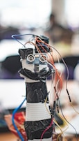 A homemade robot constructed from household materials and electronic components including a microcontroller board and several colorful wires protruding from its top. The body appears to be made from stacked containers wrapped in black tape, with a pair of ultrasonic sensors resembling eyes.