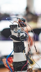 A homemade robot constructed from household materials and electronic components including a microcontroller board and several colorful wires protruding from its top. The body appears to be made from stacked containers wrapped in black tape, with a pair of ultrasonic sensors resembling eyes.