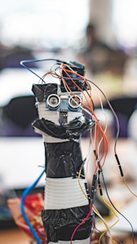 A homemade robot constructed from household materials and electronic components including a microcontroller board and several colorful wires protruding from its top. The body appears to be made from stacked containers wrapped in black tape, with a pair of ultrasonic sensors resembling eyes.
