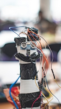 A homemade robot constructed from household materials and electronic components including a microcontroller board and several colorful wires protruding from its top. The body appears to be made from stacked containers wrapped in black tape, with a pair of ultrasonic sensors resembling eyes.