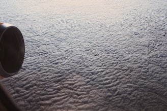 Aerial view of a dense layer of clouds with a visible airplane engine on the left. The clouds appear fluffy and textured, creating a quilt-like pattern across the horizon.