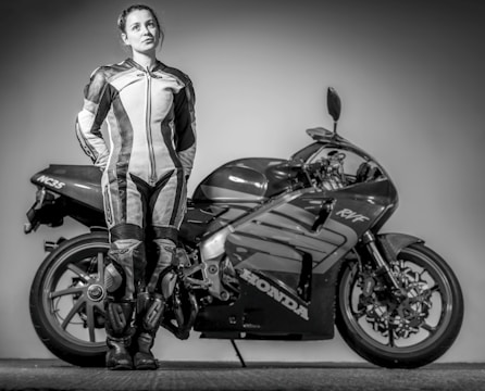 Model wearing a black motorbike suit standing beside a vintage motorcycle.