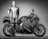 A person dressed in a full motorcycle racing suit is standing next to a sportbike. The motorcycle is a Honda model, with visible branding on the body. The image is in black and white, and the lighting highlights the contours of both the person and the bike.