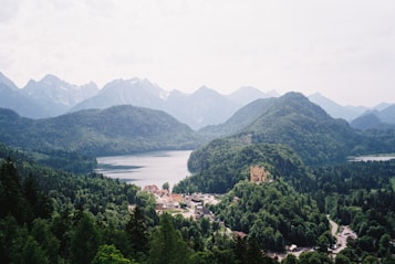 A lush green landscape featuring dense forests and rolling hills with a serene lake nestled in the valley. A historic castle perched on a hill overlooks a small village with terracotta roofs. The distant backdrop is dominated by majestic, snow-capped mountains under a soft, diffused sky.