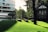 A modern, curved glass building stands on the left, next to a traditional brick house with white-framed windows on the right. The foreground features a sunny, well-manicured grassy hill with distinctive dark stone benches casting long shadows. Tall trees and lush greenery surround the area, giving a tranquil, park-like ambiance.