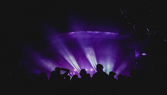 A vibrant concert scene with multicolored lights casting across a stage filled with people. Silhouettes of the audience are visible, creating a dynamic and lively atmosphere.
