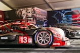 A race car with the number 13, featuring prominent branding such as Motul and Michelin, is displayed in a tent-like setting. The car is red with some black elements and is positioned next to another sleek, black sports car. The background includes promotional posters and product displays related to automotive fluids.