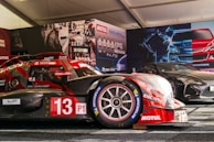A race car with the number 13, featuring prominent branding such as Motul and Michelin, is displayed in a tent-like setting. The car is red with some black elements and is positioned next to another sleek, black sports car. The background includes promotional posters and product displays related to automotive fluids.