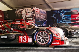 A race car with the number 13, featuring prominent branding such as Motul and Michelin, is displayed in a tent-like setting. The car is red with some black elements and is positioned next to another sleek, black sports car. The background includes promotional posters and product displays related to automotive fluids.