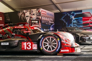 A race car with the number 13, featuring prominent branding such as Motul and Michelin, is displayed in a tent-like setting. The car is red with some black elements and is positioned next to another sleek, black sports car. The background includes promotional posters and product displays related to automotive fluids.