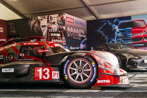 A race car with the number 13, featuring prominent branding such as Motul and Michelin, is displayed in a tent-like setting. The car is red with some black elements and is positioned next to another sleek, black sports car. The background includes promotional posters and product displays related to automotive fluids.