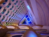 An intricate interior architecture design featuring a series of interlocking geometric patterns. The ceiling displays layers of semi-circular shapes in pastel colors, accented by green and blue lighting elements. The space is lit with ambient lighting, enhancing the modern and futuristic aesthetic.