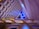 An intricate interior architecture design featuring a series of interlocking geometric patterns. The ceiling displays layers of semi-circular shapes in pastel colors, accented by green and blue lighting elements. The space is lit with ambient lighting, enhancing the modern and futuristic aesthetic.