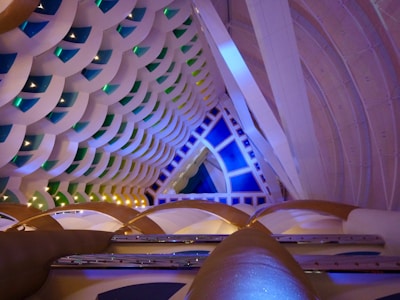 An intricate interior architecture design featuring a series of interlocking geometric patterns. The ceiling displays layers of semi-circular shapes in pastel colors, accented by green and blue lighting elements. The space is lit with ambient lighting, enhancing the modern and futuristic aesthetic.