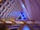 An intricate interior architecture design featuring a series of interlocking geometric patterns. The ceiling displays layers of semi-circular shapes in pastel colors, accented by green and blue lighting elements. The space is lit with ambient lighting, enhancing the modern and futuristic aesthetic.