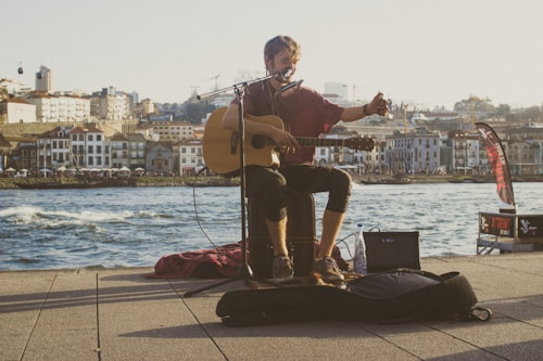A street musician plays an acoustic guitar while sitting by a waterfront. The sun sets in the background, casting a warm glow over the scene. A small amplifier and a microphone are set up in front of him. Buildings and a hilly landscape are visible across the water, and the overall atmosphere is relaxed and casual.