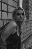 A young woman with blonde hair is leaning against a textured brick wall. She is wearing a dark tank top and a denim jacket that is slipping off her shoulders. Her expression is neutral, and she is gazing directly at the camera. The image is in black and white, adding a dramatic and timeless quality.