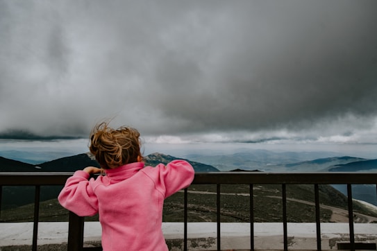 A young child wearing a bright pink jacket stands facing a railing, looking out over a vast mountainous landscape under a cloudy sky. The scene suggests a sense of wonder and exploration.