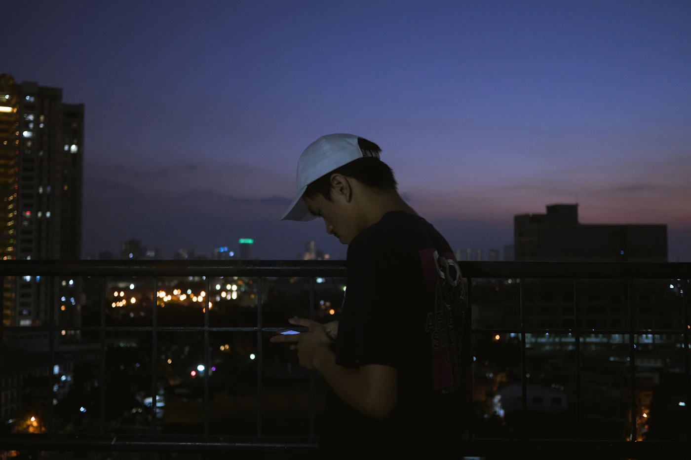 A man stands alone on a balcony at night, looking at his phone, city lights stretching out behind him